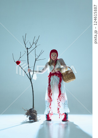 Woman in white-red layered dress stands beside uprooted tree, woven basket in hand, evoking symbolism of displaced growth and transformation. 124615887