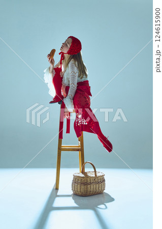 Young woman in white lace dress and red accents reclining on stool, lifting bread to lips, merging hunger with seduction. 124615900
