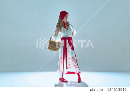 Young woman in white lace dress with red accents, evoking fairytale imagery, gracefully posing with basket, symbolizing contrast between innocence and power. 124615911