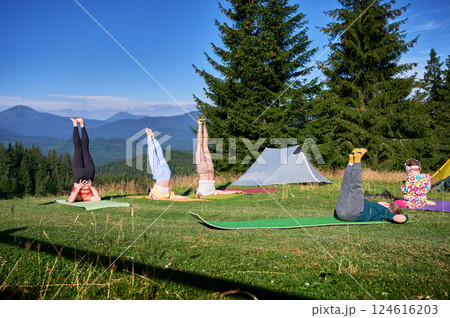 Group of people doing yoga pose outdoor in camping in the mountains. Adults and children on yoga mats, each doing a yoga pose under a clear blue sky in the morning. Young boy is instructor. 124616203