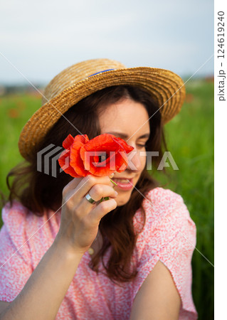 A young woman in a straw hat holds a vibrant red poppy flower near her face in a sunny field A young woman in a straw hat holds a vibrant red poppy flower near her face in a sunny field 124619240