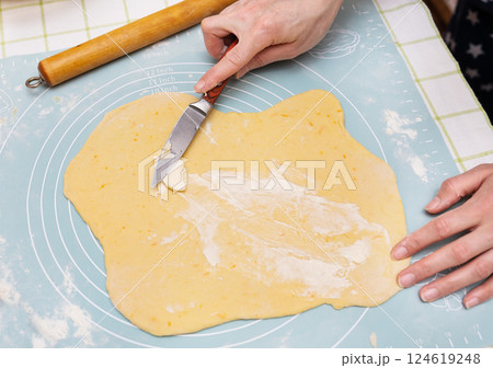 A young Caucasian female baker spreads butter on sweet dough, preparing Easter bread. She uses a knife to evenly distribute the filling. Preparation for a delicious holiday treat 124619248