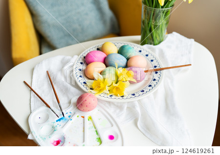 Pastel Easter eggs, daffodils, and paintbrushes are artfully arranged on a white table. This image evokes spring and the joyful celebration of Easter 124619261