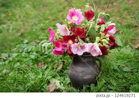 A rustic dark vase holds a vibrant bouquet of pink and crimson hollyhocks, nestled amidst greenery A rustic dark vase holds a vibrant bouquet of pink and crimson hollyhocks, nestled amidst greenery 124619271
