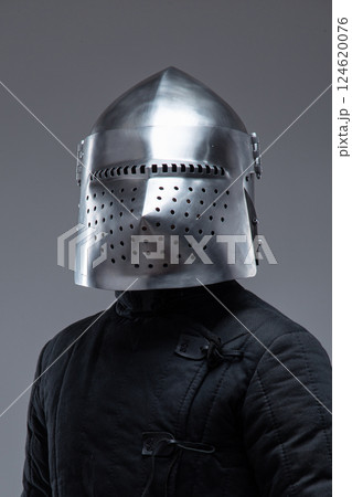 Close-Up of a Medieval Knight's Helmet with Perforated Visor. This image focuses on a shiny metal helmet with numerous small holes in the visor, worn by a person in a dark quilted garment. Close-Up of a Medieval Knight's Helmet with Perforated Visor. This image focuses on a shiny metal helmet with numerous small holes in the visor, worn by a person in a dark quilted garment. 124620076