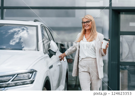 Outdoors with cup of drink. Woman in white formal clothes is in the car dealership Outdoors with cup of drink. Woman in white formal clothes is in the car dealership 124623353