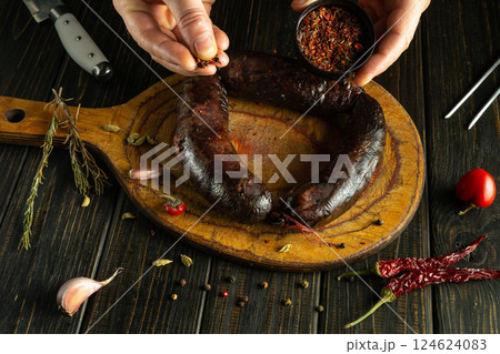 A pair of hands adds seasoning to a dark sausage resting on a wooden board. Surrounding ingredients include garlic, herbs, and chili, enhancing the culinary atmosphere 124624083