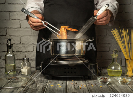 In a modern kitchen, a chef skillfully uses a fork and knife to handle the chicken legs cooking in a pan. The warm atmosphere is illuminated by the stove In a modern kitchen, a chef skillfully uses a fork and knife to handle the chicken legs cooking in a pan. The warm atmosphere is illuminated by the stove 124624089