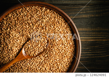 A wooden bowl filled with golden buckwheat grains sits on a dark wooden surface. A wooden spoon rests on the edge, highlighting the natural beauty and texture of the seeds. 124624091