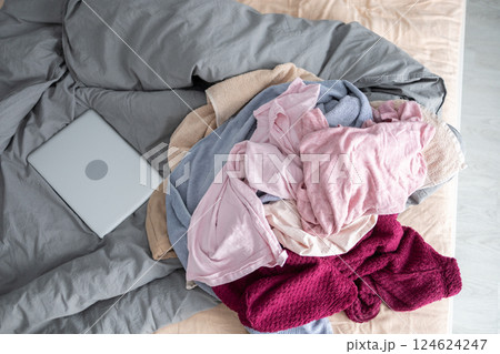 Messy pile of clothes and laptop on unmade bed in bedroom. Household clutter and everyday disorganization in a lived-in home. 124624247