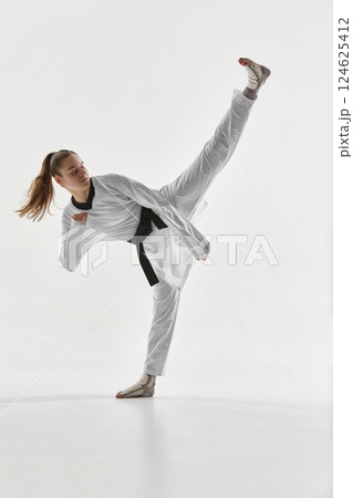 Young woman, taekwondo athlete performing high kick in studio showcasing strength balance and flexibility. 124625412