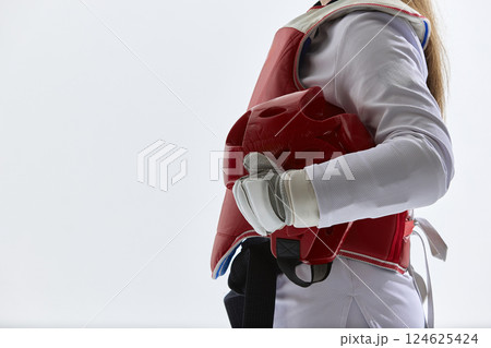 Cropped image of female taekwondo fighter standing in red protective gear, holding helmet isolated on white background. 124625424