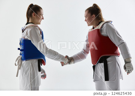 Two smiling women, taekwondo athletes in red and blue gear shaking hands after match in sign of mutual respect. Two smiling women, taekwondo athletes in red and blue gear shaking hands after match in sign of mutual respect. 124625430