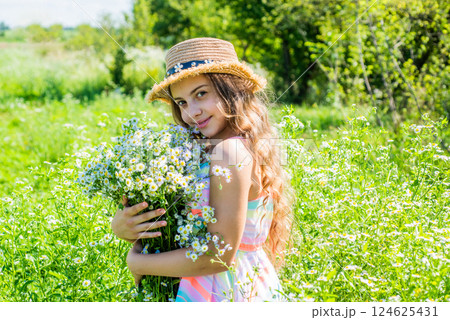 Happy little kid wear sun hat on long wavy hair holding beautiful chamomile flowers spa and beauty care on sunny summer landscape, look 124625431