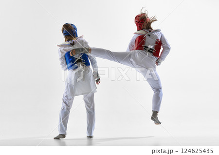 Woman in red gear jumping mid-air to strike blue female opponent with powerful side kick during match on white background. Woman in red gear jumping mid-air to strike blue female opponent with powerful side kick during match on white background. 124625435