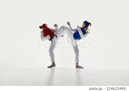 Female taekwondo fighters in red and blue protective gear in motion, fighting with synchronized kicks on white background. 124625468