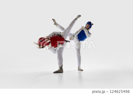 Female fighter in red executing high side kick at opponent in blue gear during combat against white studio background. 124625498
