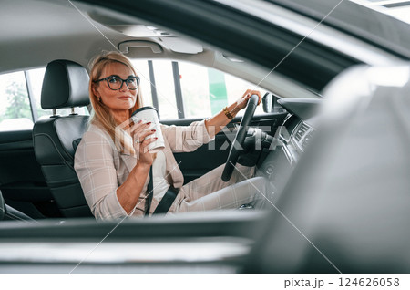 Driving vehicle and holding cup of drink. Woman in white formal clothes is in the car dealership Driving vehicle and holding cup of drink. Woman in white formal clothes is in the car dealership 124626058