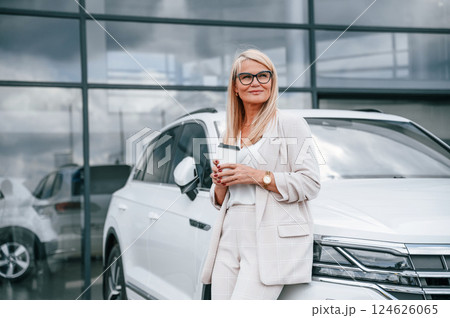 Outside with cup of drink. Woman in white formal clothes is in the car dealership Outside with cup of drink. Woman in white formal clothes is in the car dealership 124626065