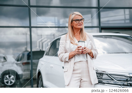 Outside with cup of drink. Woman in white formal clothes is in the car dealership Outside with cup of drink. Woman in white formal clothes is in the car dealership 124626066