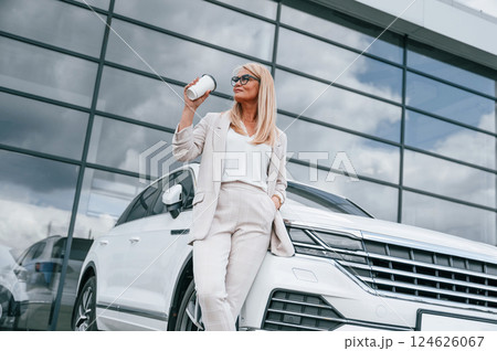 Outside with cup of drink. Woman in white formal clothes is in the car dealership Outside with cup of drink. Woman in white formal clothes is in the car dealership 124626067