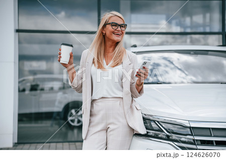Standing against building with big windows. Woman in white formal clothes is in the car dealership 124626070