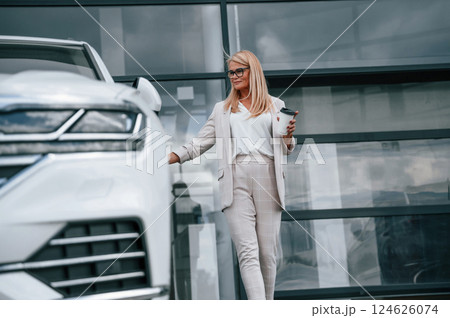 Standing outdoors against building and holding drink. Woman in white formal clothes is in the car dealership 124626074