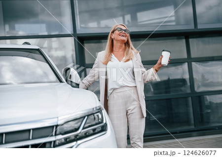 Standing outdoors against building and holding drink. Woman in white formal clothes is in the car dealership 124626077