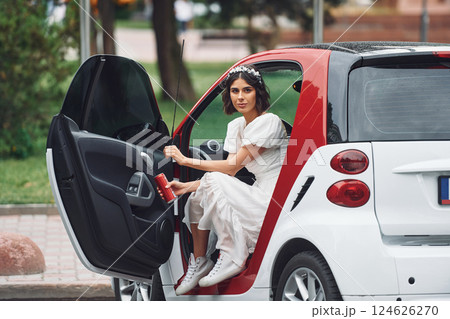 Sitting in white and red colored car. Bride in dress is outdoors before her wedding begins Sitting in white and red colored car. Bride in dress is outdoors before her wedding begins 124626270