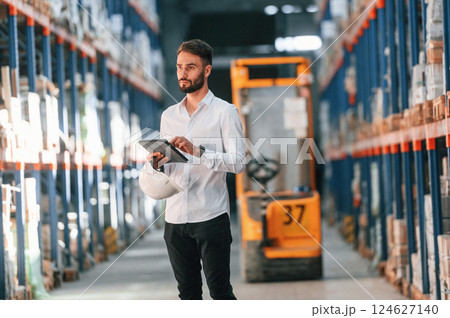 Standing and holding tablet in hands. Storage worker is in the warehouse with bunch of products Standing and holding tablet in hands. Storage worker is in the warehouse with bunch of products 124627140
