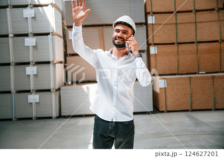 Talking by phone and greeting with someone. Storage worker is in the warehouse with bunch of products 124627201
