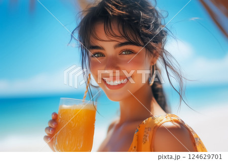 Portrait of beautiful young caucasian woman on beach against background of blue sky holding glass of orange juice and smiling looking at camera 124629701