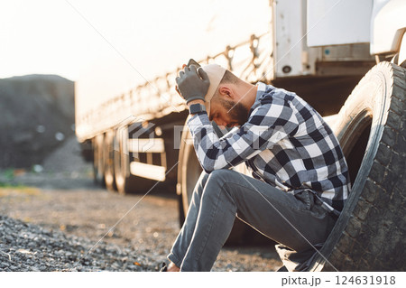Sitting with phone, waiting for help. Damaged tire. Young truck driver is with his vehicle at daytime Sitting with phone, waiting for help. Damaged tire. Young truck driver is with his vehicle at daytime 124631918