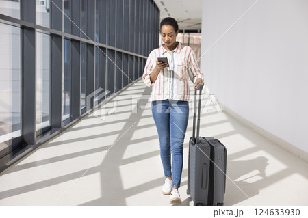 Woman rolling suitcase, using smartphone while walking through modern hallway Woman rolling suitcase, using smartphone while walking through modern hallway 124633930