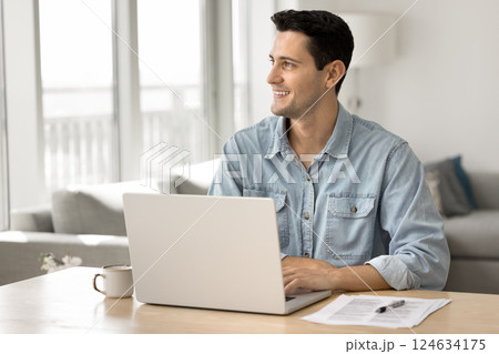 Young man sitting at desk with laptop, smiling, looking away 124634175