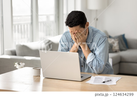 Man covers face with hands seated at desk with laptop 124634299