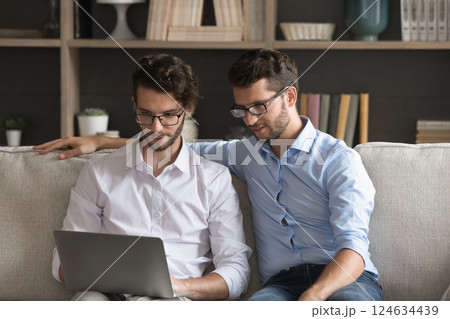 Friendly young twin brothers sit on couch use notebook together Friendly young twin brothers sit on couch use notebook together 124634439