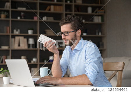 Busy gen z male sitting by notebook using phone loudspeaker Busy gen z male sitting by notebook using phone loudspeaker 124634463