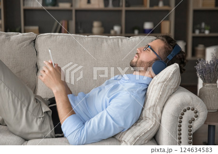 Young man wearing wireless headphones relaxing on couch holding cellphone 124634583