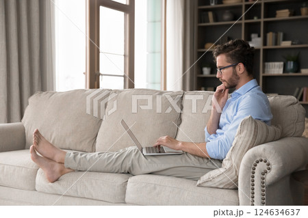 Pensive millennial man sitting on sofa holding notebook on laps Pensive millennial man sitting on sofa holding notebook on laps 124634637