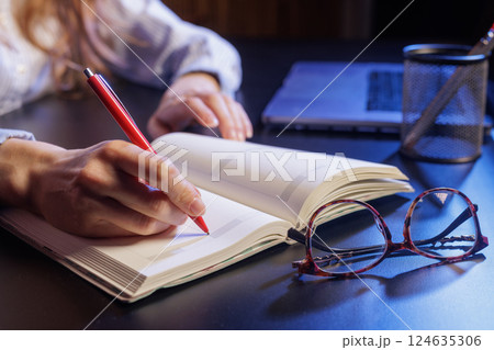 A person writing in a notebook with a red pen, a laptop and a pen holder in the background. Eyeglasses are placed on the table next to the notebook. A person writing in a notebook with a red pen, a laptop and a pen holder in the background. Eyeglasses are placed on the table next to the notebook. 124635306