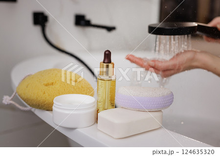 Spa products and skincare essentials on bath edge, hand under shower water in modern bathroom, self-care concept. 124635320