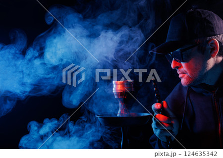 Man smoking hookah, adjusting hot charcoal with tongs in a smoky, moody atmosphere, illuminated by red and blue lighting, 124635342
