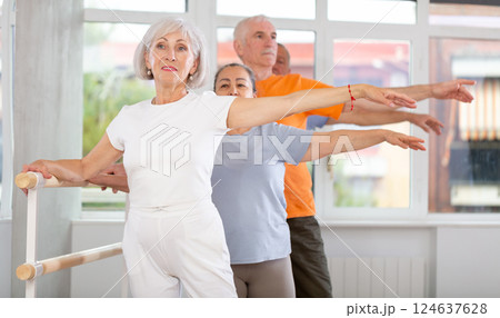 Dancing positive elderly women and men engaged in group class perform exercise near ballet barre, standing in 2nd position of ballet stance in dance studio 124637628