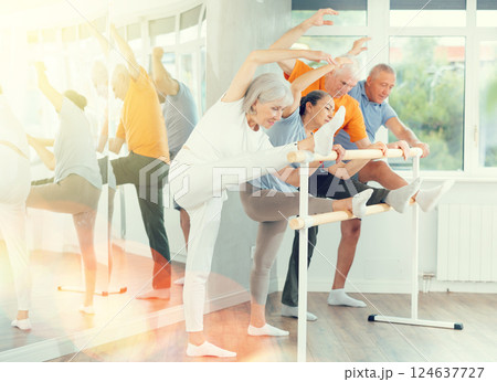 Active mature woman practicing leg stretching holding by ballet barre together with others 124637727