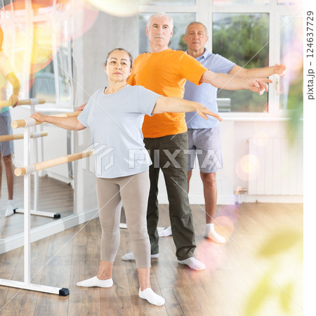 Dancing positive elderly women and men engaged in group class perform exercise near ballet barre, standing in 2nd position of ballet stance in dance studio 124637729
