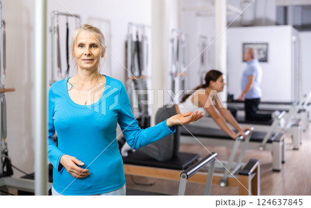 Portrait of smiling aged woman in activewear posing against people exercising on reformer bed machine in Pilates studio Portrait of smiling aged woman in activewear posing against people exercising on reformer bed machine in Pilates studio 124637845