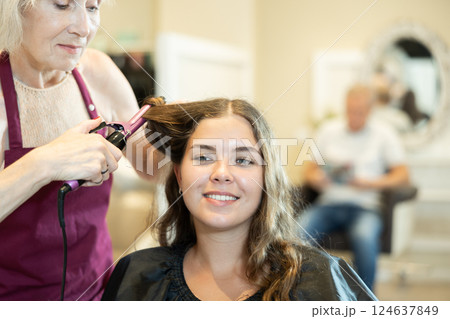 Female hairdresser makes curls the hair of young woman using curling iron Female hairdresser makes curls the hair of young woman using curling iron 124637849