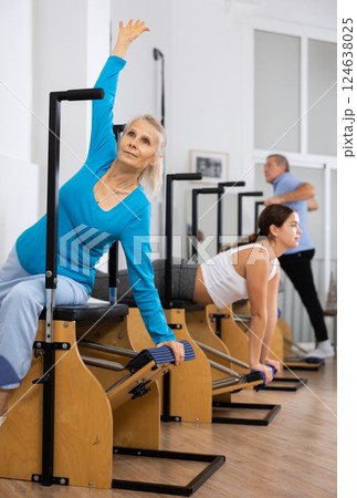 Aged female in activewear concentrating on stretching exercise on Pilates chair equipment in gym. Active and healthy lifestyle rehabilitation concept 124638025