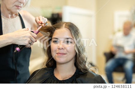 Experienced hairdresser using curling wands to create waves to young woman Experienced hairdresser using curling wands to create waves to young woman 124638111